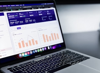 A laptop with an open screen displaying dashboard analytics featuring charts, graphs, and numerical data related to sales and subscriptions. The interface is predominantly dark-themed with sections highlighting key performance metrics. The device shown is a MacBook Pro, indicated by the keyboard layout and branding.