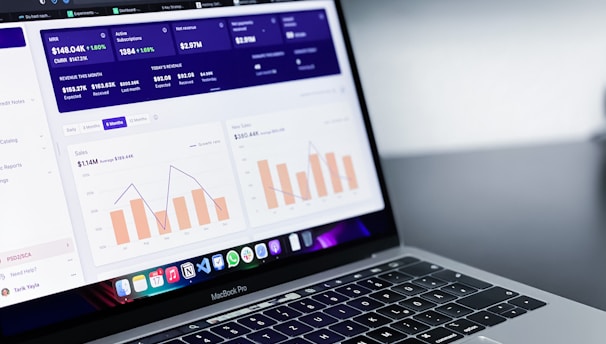 A laptop with an open screen displaying dashboard analytics featuring charts, graphs, and numerical data related to sales and subscriptions. The interface is predominantly dark-themed with sections highlighting key performance metrics. The device shown is a MacBook Pro, indicated by the keyboard layout and branding.